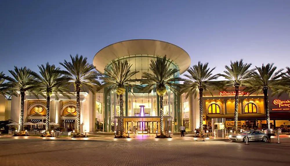 The image shows a dusk view of a stylish modern shopping mall entrance flanked by palm trees and featuring upscale storefronts