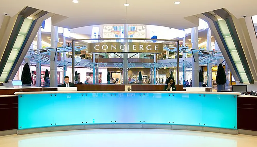 A modern concierge desk with two staff members is situated centrally in a brightly lit mall with escalators in the background