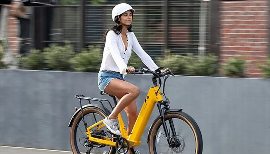 A person is riding a yellow electric bicycle on the street, wearing a white helmet and casual clothing.
