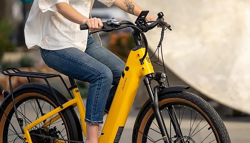 A person is seated on a yellow electric bicycle with a rear cargo rack holding the handlebars and appears ready to ride