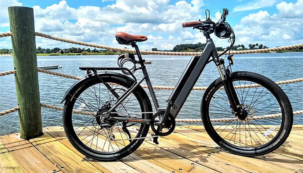 A modern electric bicycle is parked on a wooden pier overlooking a scenic lake bordered by rope railings and greenery on a sunny day