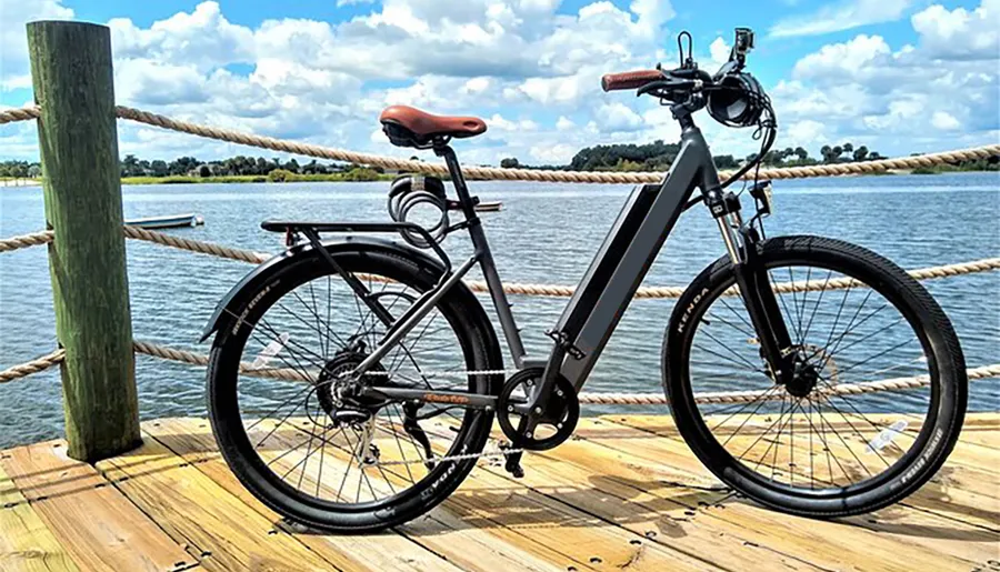 A modern electric bicycle is parked on a wooden pier overlooking a scenic lake bordered by rope railings and greenery on a sunny day.