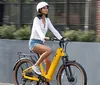 A person is riding a yellow electric bike on a city street while wearing a white helmet a white shirt and denim shorts