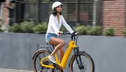 A person is riding a yellow electric bike on a city street while wearing a white helmet, a white shirt, and denim shorts.