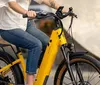 A person is riding a yellow electric bike on a city street while wearing a white helmet a white shirt and denim shorts