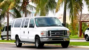 A white Ford passenger van is driving on a road lined with palm trees.