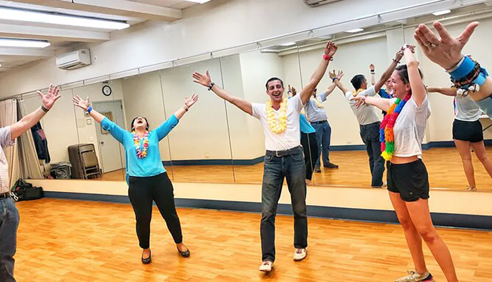 A group of people wearing leis are joyfully stretching their arms upwards in a dance studio with mirrors on the wall