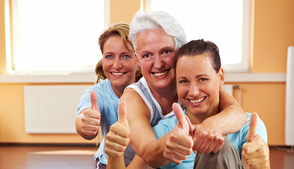 Three women in sporty attire are smiling at the camera giving a thumbs-up in a brightly lit room