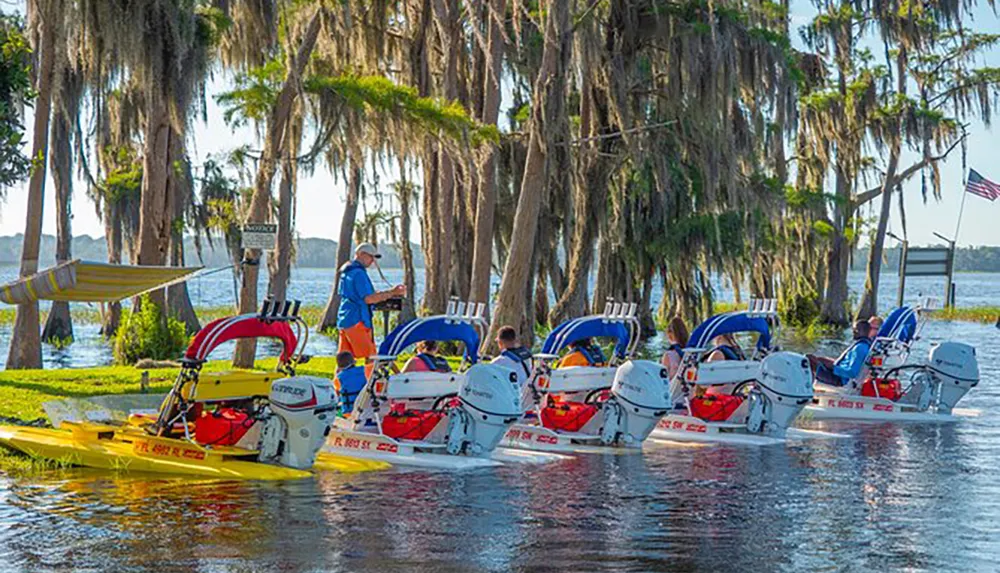 A group of people is preparing various motorboats for an outing near a body of water surrounded by Spanish moss-draped trees with an individual standing onshore to the left