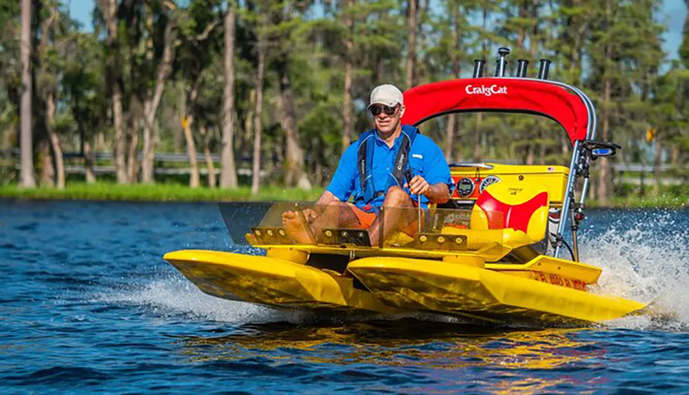 A man wearing a life jacket and sunglasses is steering a yellow compact boat with the label CraigCat through a body of water with greenery and trees in the background