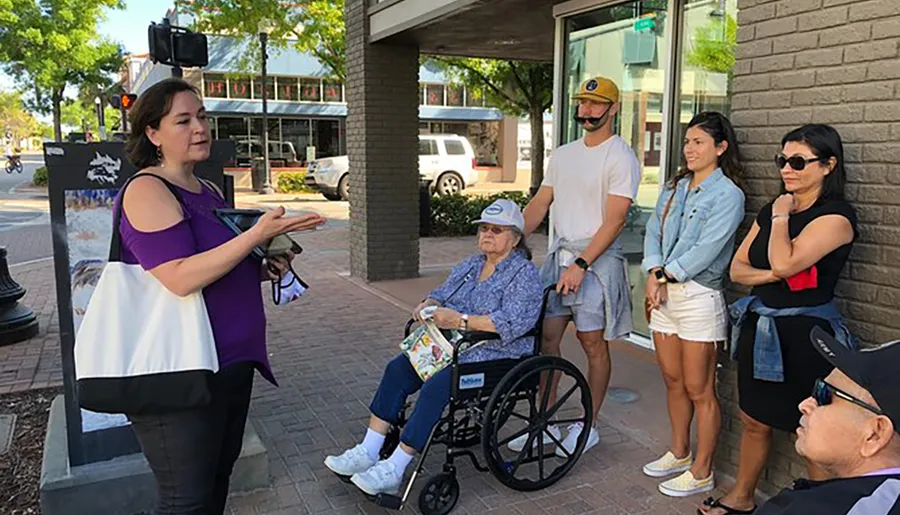 A group of people, including an individual in a wheelchair, attentively listens to a woman who appears to be giving a tour or presentation outdoors.