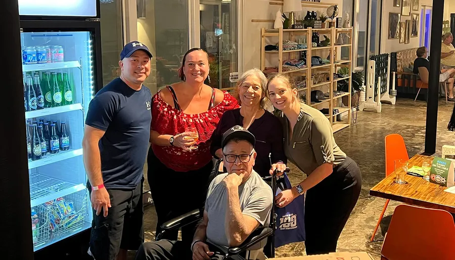 Five people are posing for a photo in a casual indoor setting, with a seated gentleman in a wheelchair in the foreground and a display fridge with drinks to the left.