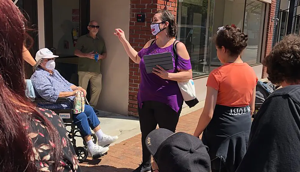 A group of people including a woman with a mask gesturing with her hand listen to someone speaking standing outside on a sunny day