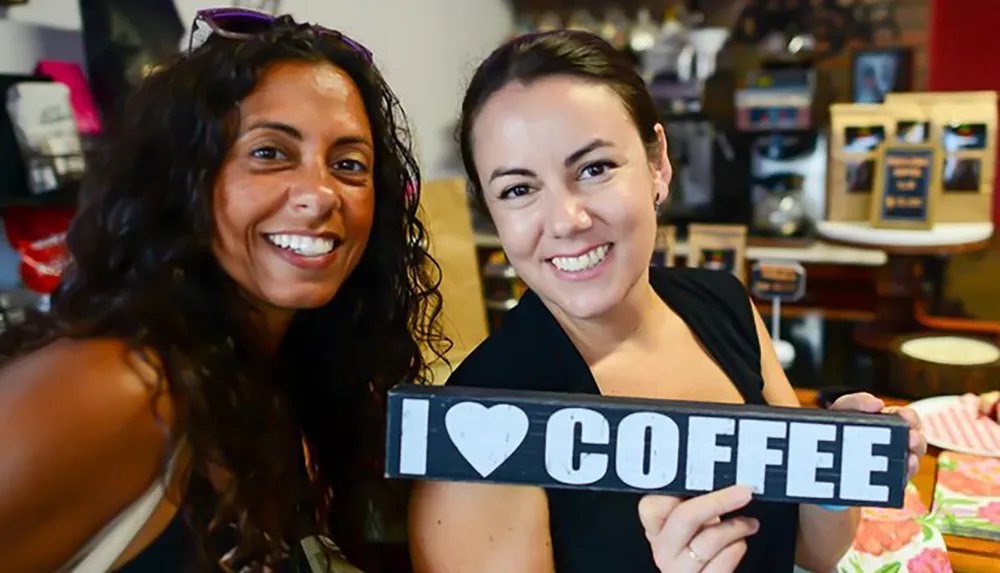 Two smiling women are holding a sign that reads I  COFFEE in a coffee shop