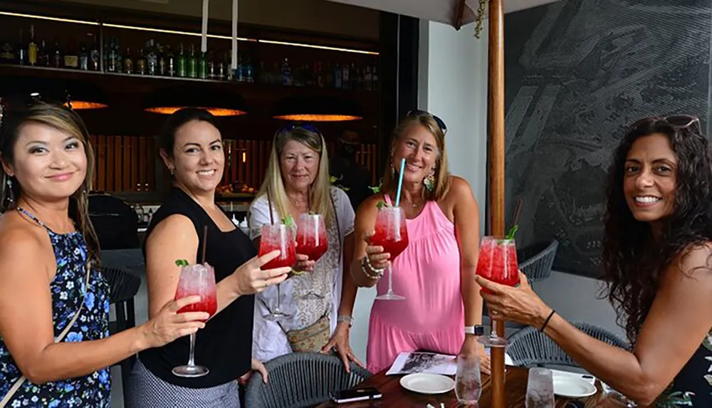 Five women are smiling and toasting with glasses of red cocktails at a bar