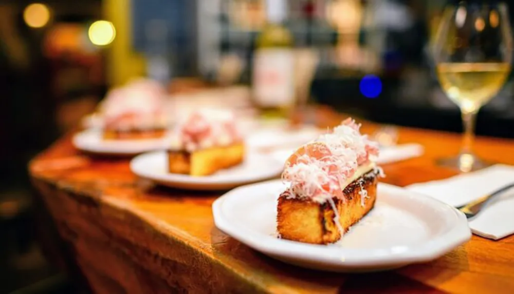 A row of gourmet toasts topped with a generous amount of shredded meat is presented on white plates along a bar with a glass of white wine in the background creating a cozy dining atmosphere