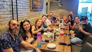 A group of people is smiling and posing for a photo while sitting at a long dining table with drinks, suggesting a social gathering or a celebration.