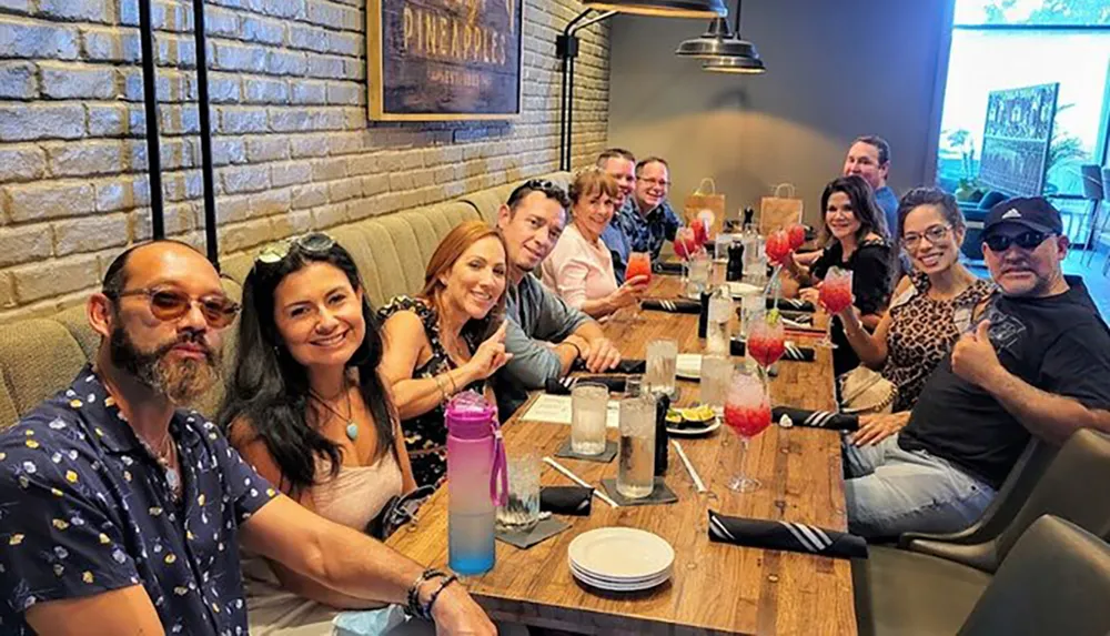A group of people is smiling and posing for a photo while sitting at a long dining table with drinks suggesting a social gathering or a celebration