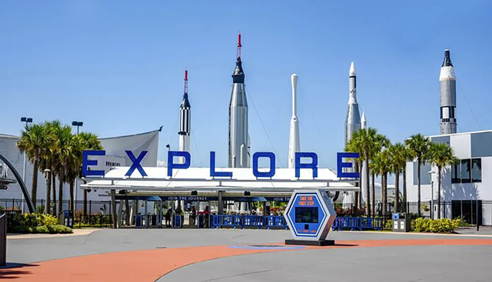 The image features the entrance to a space-themed attraction with the word EXPLORE emblazoned above and a display of historic rockets in the background under a clear blue sky