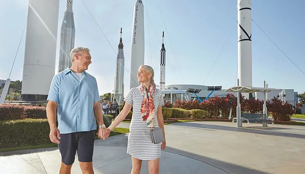 A smiling couple is walking hand in hand near several rocket displays at a space center exhibit under a clear blue sky