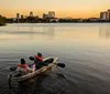 Two individuals are smiling while kayaking in an urban waterway at dusk with city lights starting to twinkle in the background