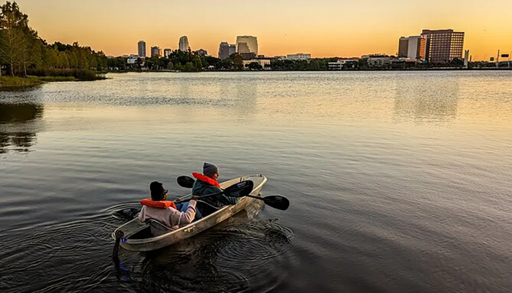 Two people in life jackets are paddling a kayak on calm waters with a city skyline in the background during sunset