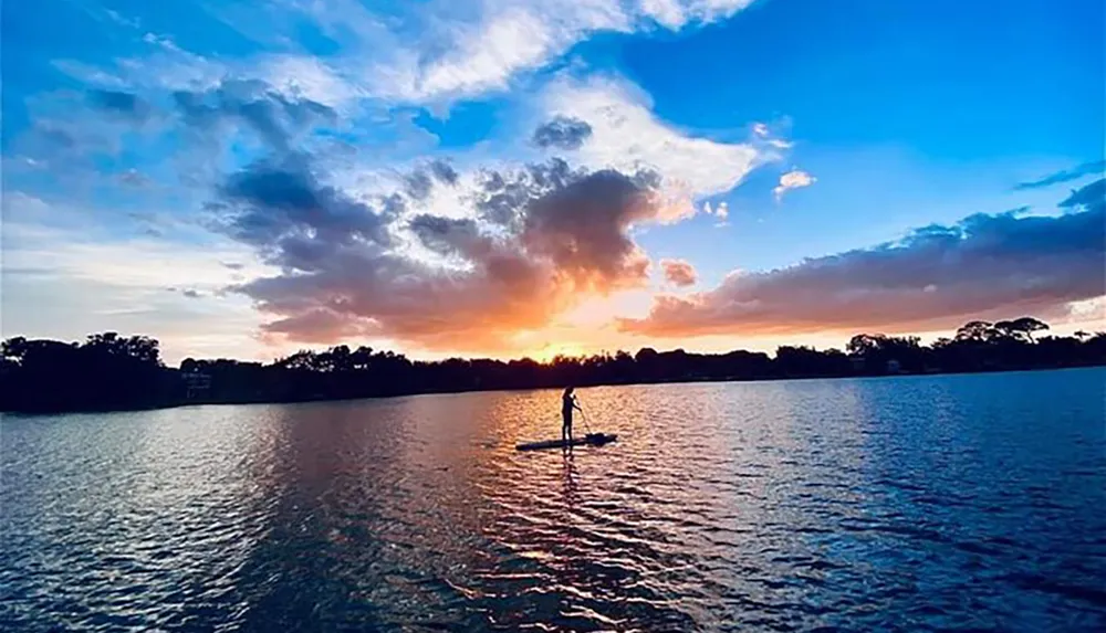 A person is paddleboarding on tranquil waters against a vivid sunset sky