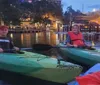 Two individuals are smiling while kayaking in an urban waterway at dusk with city lights starting to twinkle in the background