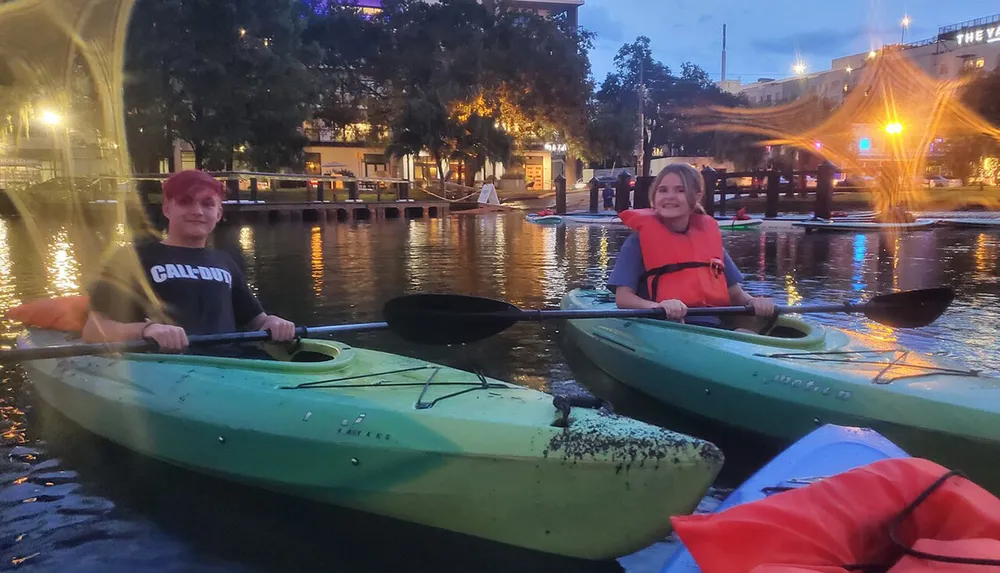 Two individuals are smiling while kayaking in an urban waterway at dusk with city lights starting to twinkle in the background