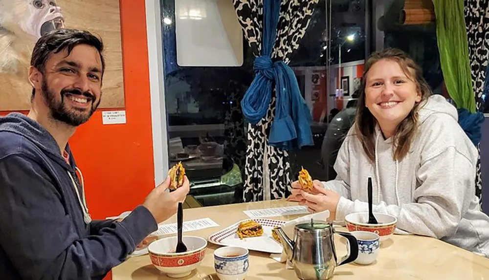 A man and a woman are smiling at the camera while holding sandwiches with bowls and a teapot on the table in front of them in a brightly colored dining area