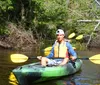 A person wearing a life jacket and cap is kayaking on a calm river surrounded by lush greenery under a clear sky