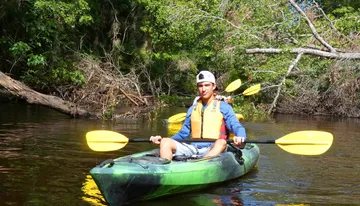 A person wearing a life jacket and cap is kayaking on a calm river surrounded by lush greenery under a clear sky.