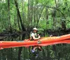 A person wearing a life jacket and a hat is paddling a bright orange kayak through calm waters surrounded by a lush green forest