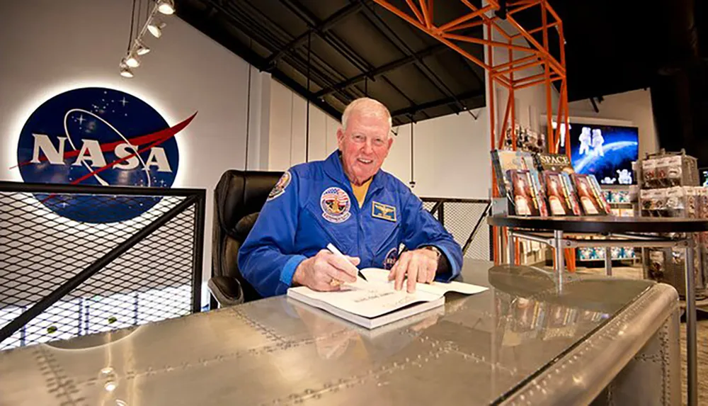 A person wearing a blue astronaut flight suit is signing autographs at a desk with a NASA emblem in the background