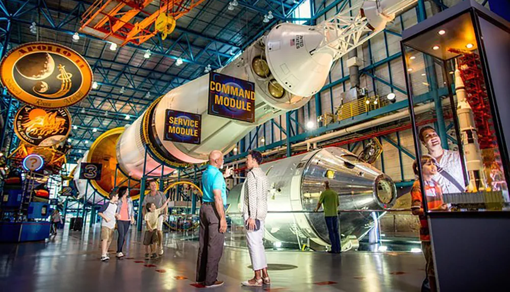 The image shows visitors exploring a space museum exhibit featuring components of a spacecraft and educational displays