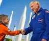 A child dressed in an orange astronaut flight suit shakes hands with an adult astronaut in a blue flight suit against a backdrop of rocket models
