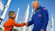 A child dressed in an orange astronaut flight suit shakes hands with an adult astronaut in a blue flight suit against a backdrop of rocket models.