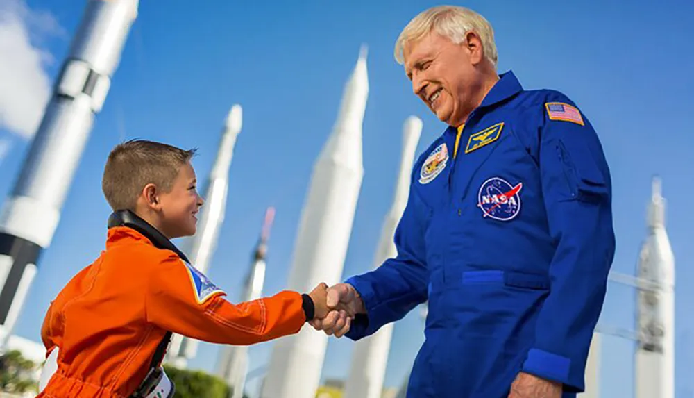 A child dressed in an orange astronaut flight suit shakes hands with an adult astronaut in a blue flight suit against a backdrop of rocket models