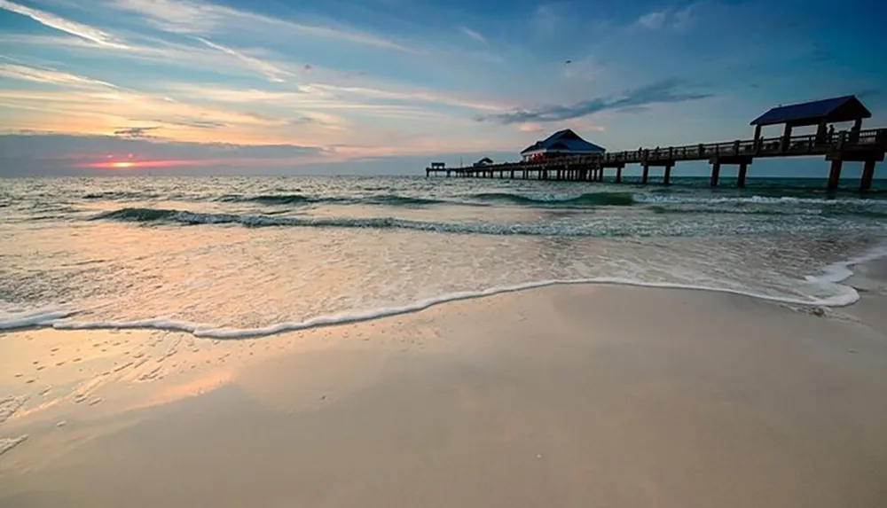 A serene sunset paints the sky with warm hues as gentle waves kiss a sandy shore with a wooden pier extending into the sea