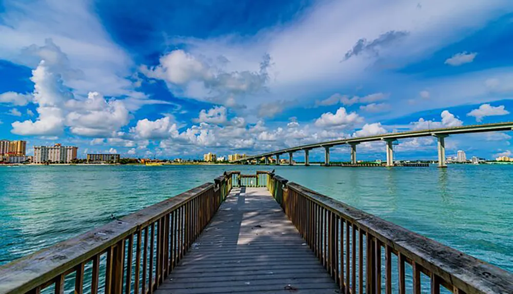 A wooden pier extends towards a body of water with a striking blue sky above and a city skyline in the distance underscored by an elevated bridge parallel to the pier