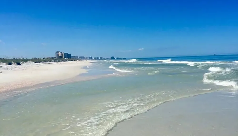 The image shows a tranquil beach with clear blue skies white sand gentle waves and a distant skyline of buildings along the coast