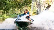 Two individuals are enjoying a ride on a jet ski, creating a splash of water behind them.