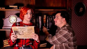 A woman and a man are in a dimly lit, vintage-style room, where the woman appears concerned while holding a newspaper with the headline 