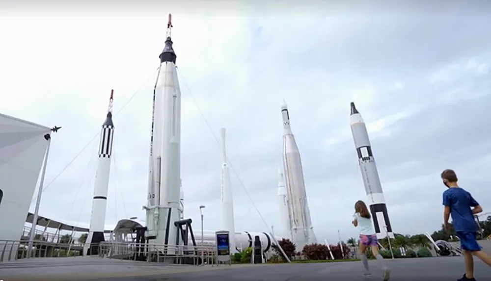 The image shows an outdoor display of various historic rockets with two children running towards them likely at a space museum or center