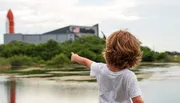 A child with curly hair, seen from behind, is pointing towards a rocket or space shuttle displayed near a building with 