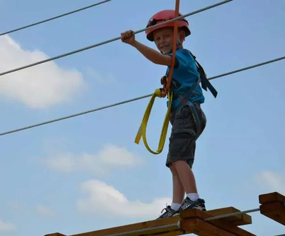 A child wearing a helmet and safety harness is balancing on a high ropes course