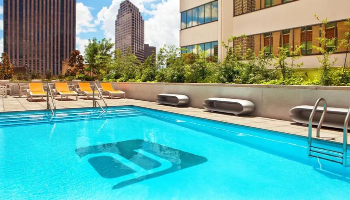 A rooftop swimming pool surrounded by lounge chairs basks in sunlight amidst urban skyscrapers