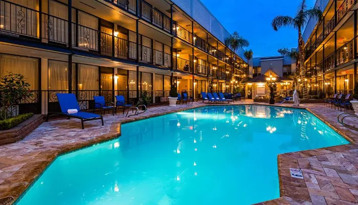 The image shows an inviting hotel pool area at twilight surrounded by rooms with balconies that are warmly lit against the evening sky