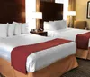 The image shows a neatly arranged hotel room with two queen-sized beds adorned with white linens and red bed runners complemented by wooden headboards bedside tables lamps and wall art