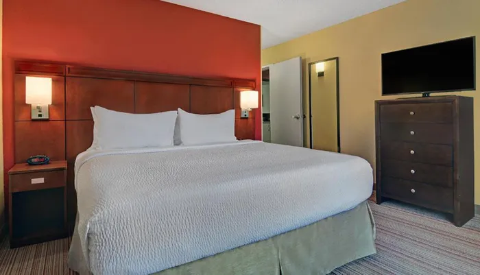 The image shows a neat and simply furnished hotel room with a large bed nightstand and a mounted television opposite the bed